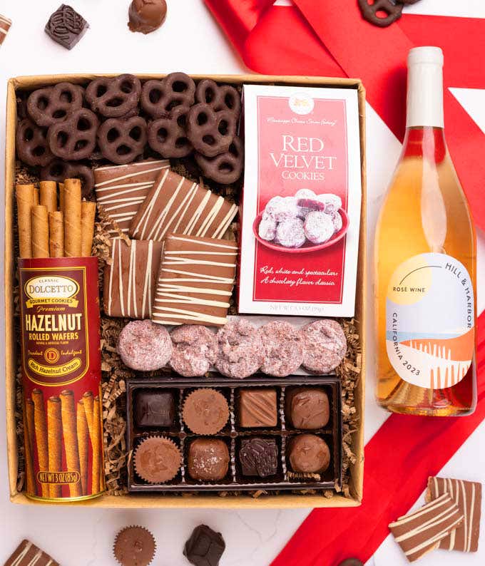 A gift box with chocolate pretzels, chocolate graham crackers, red velvet cookies, hazelnut wafer rolls, an 8 piece box of chocolates and a bottle of rosé wine.